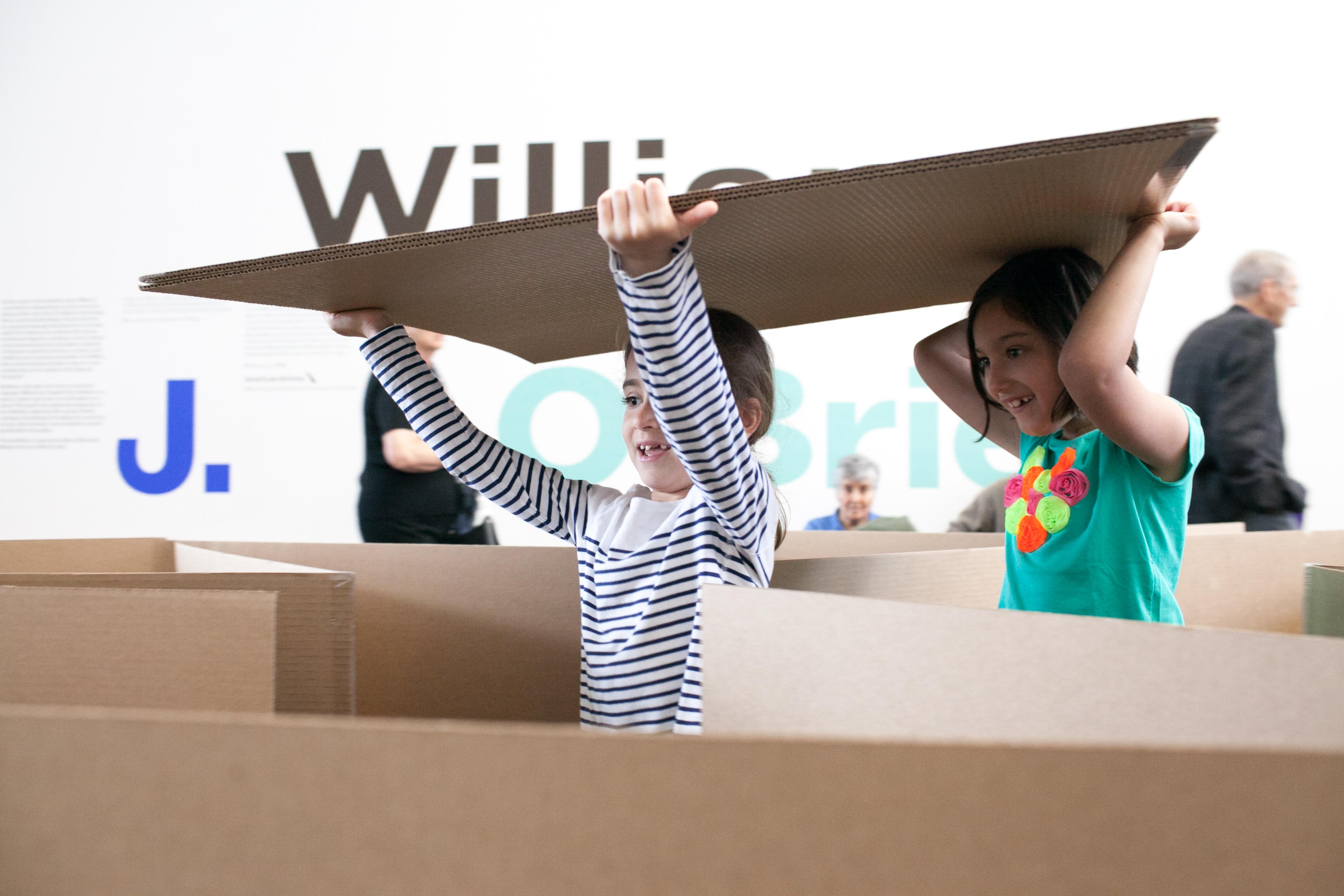 Two girls carry a sheet of cardboard above their heads as they wander through a cardboard maze. Their eyes are filled with wonder as they explore the maze.