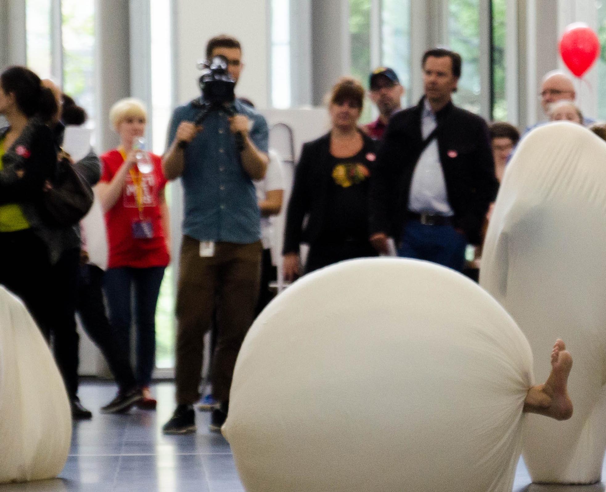 Visitors gather around performers in large cloth spheres while a cameraman films the scene.